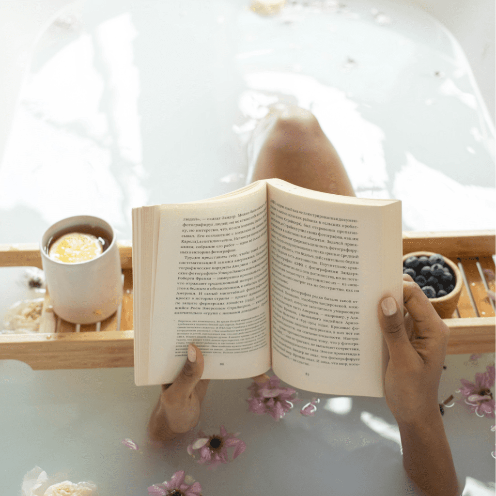 A women reading a book with tea and blueberries in a bath filled with flower petals.