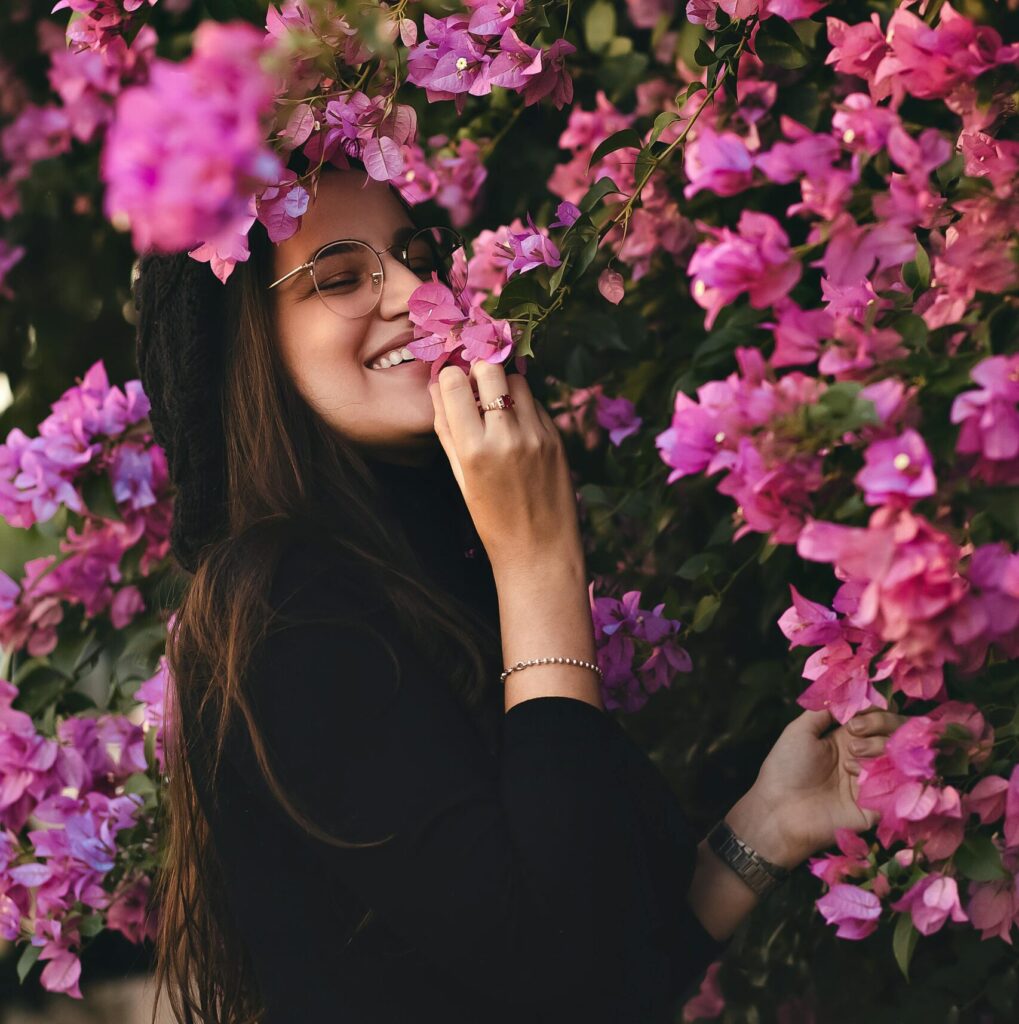 Happy, smiling women with glasses smelling pink flowers that are surrounding her.