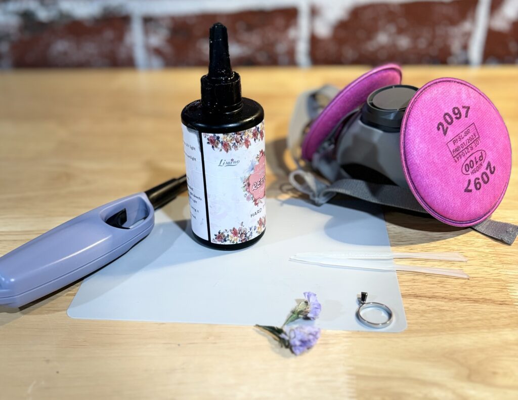 Pictured are the 7 tools recommended for resin jewelry making: resin, jewelry findings/bezels, fillers (such as dried flowers), silicon mixing sticks, a lighter, safety supplies (gloves & respirator), and protection for your surface like a silicon mat.